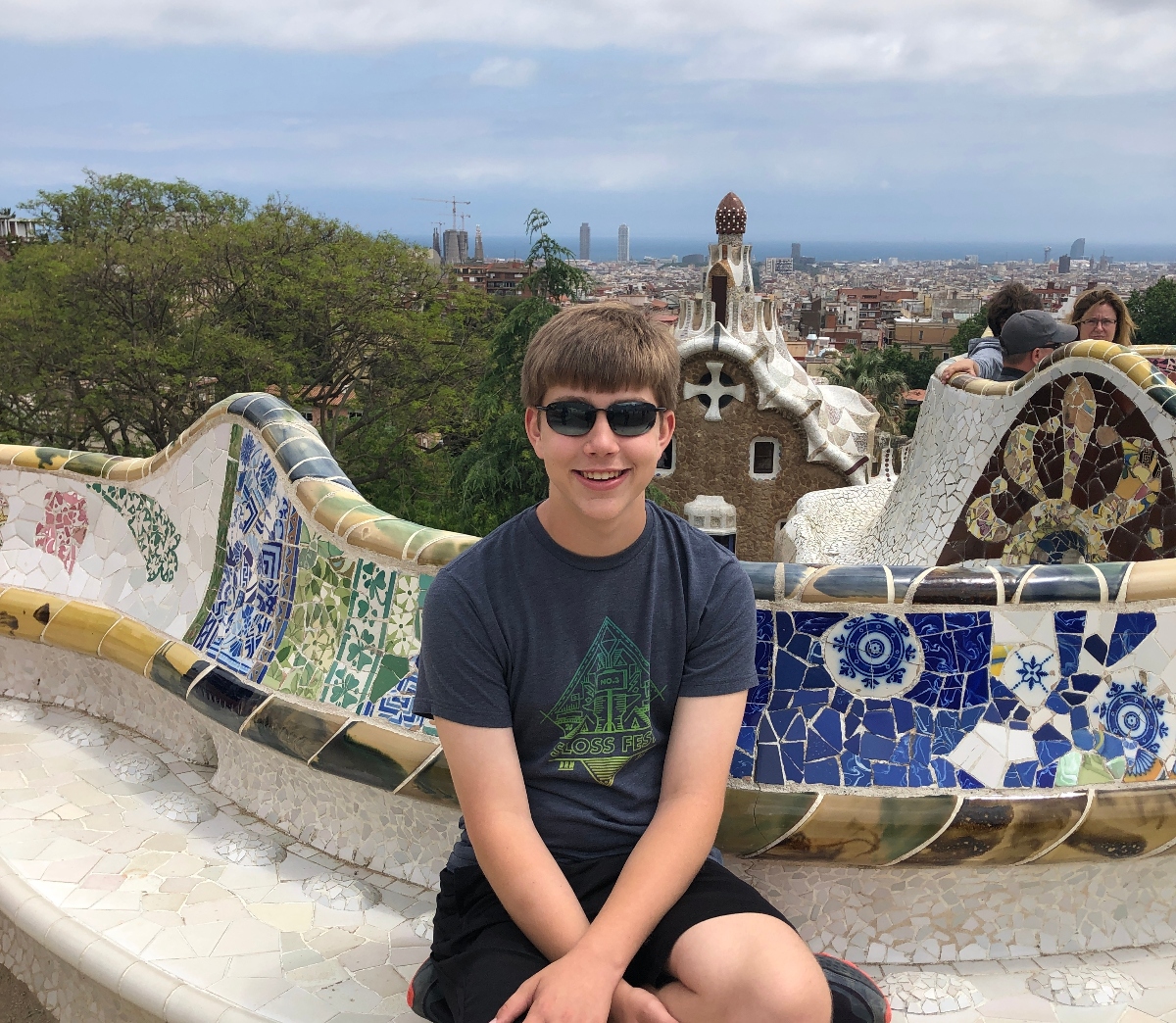 A teen boy wearing sunglasses and a dark t-shirt sits on a curved mosaic bench featuring colorful ceramic tiles in blue, green, and white patterns. The setting is an elevated viewpoint overlooking a city skyline, with trees and other visitors visible in the background.