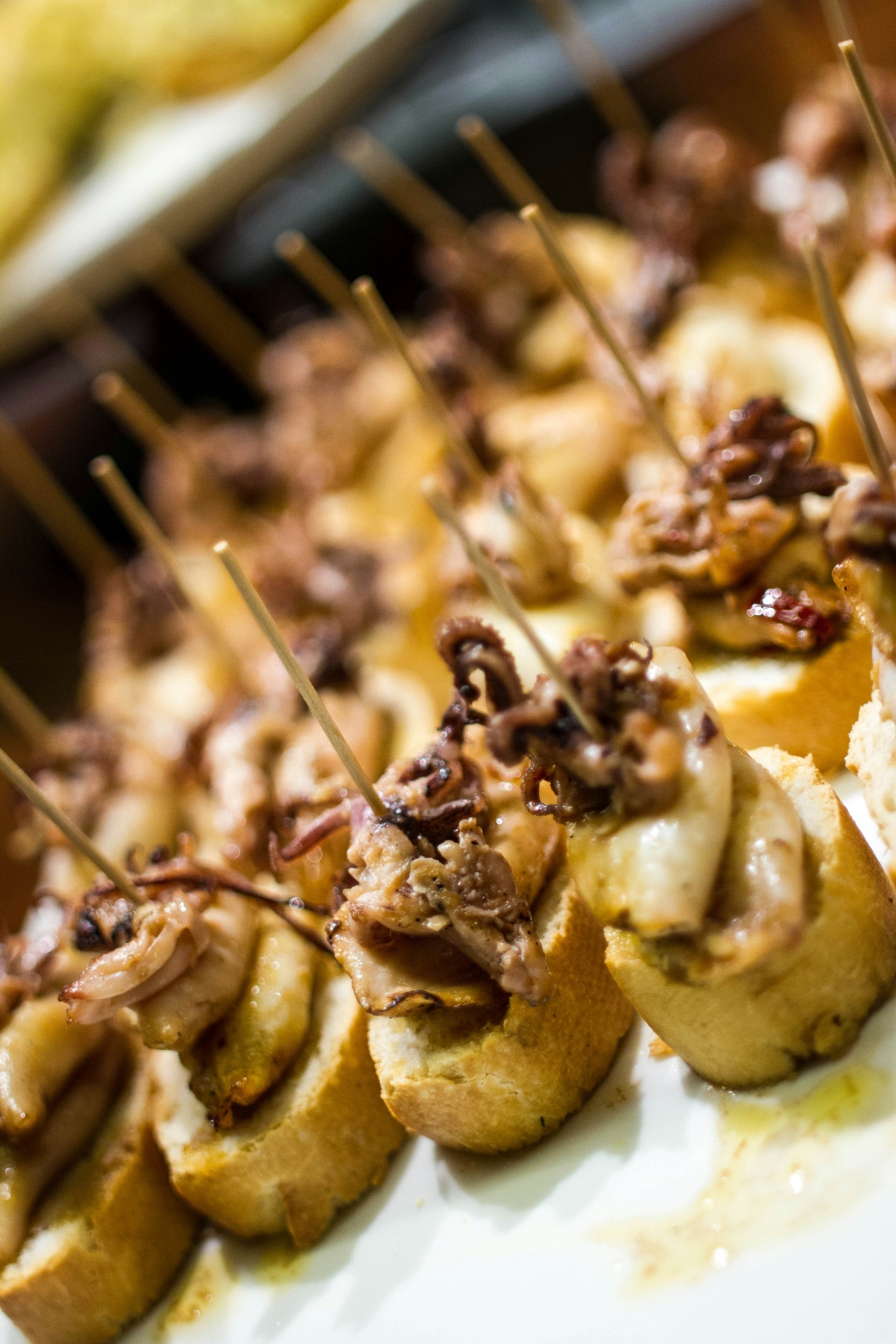 Small appetizers arranged on a white serving platter feature golden-brown bread slices topped with cooked octopus and what appear to be mushrooms or potatoes, each secured with a wooden toothpick. The hors d'oeuvres are photographed in warm lighting that emphasizes their golden-brown colors and creates a shallow depth of field effect.