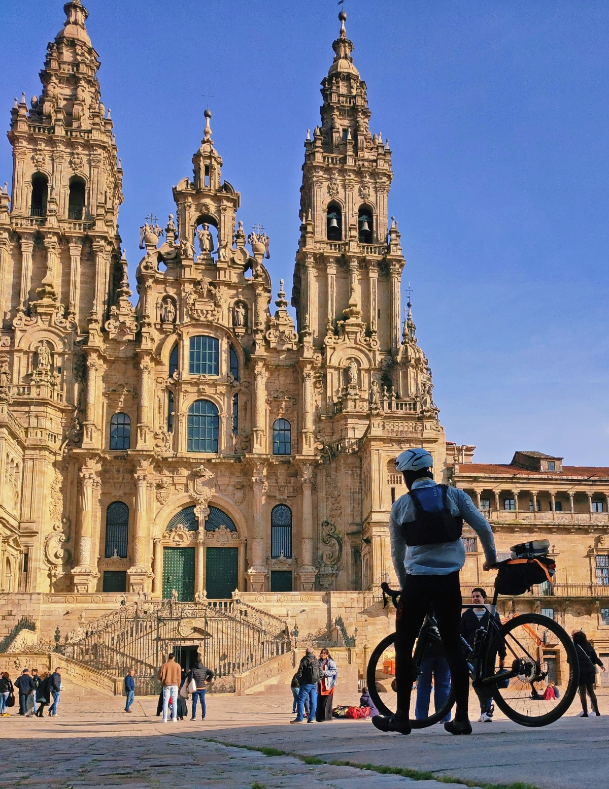 A person with a bicycle stands in the foreground of a large stone plaza facing an ornate cathedral with multiple baroque towers and an elaborately decorated facade featuring intricate stonework and sculptures. The cathedral's towering spires rise against a clear blue sky, while groups of visitors gather on the steps and plaza area in front of the grand entrance.