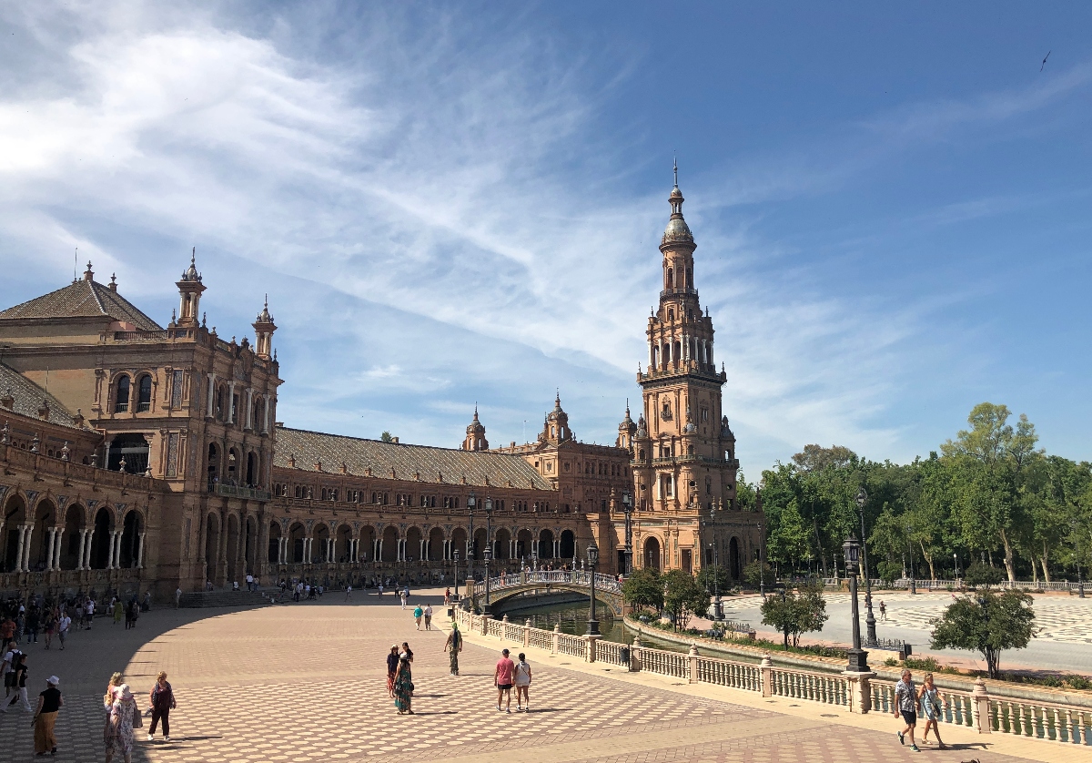 A large semicircular plaza features an ornate building complex with continuous arched galleries and multiple decorative towers, including a prominent tall spire with intricate tilework. Visitors walk across the patterned tile courtyard and over a small ornamental bridge, surrounded by Renaissance-style architecture constructed in warm-toned stone and brick.