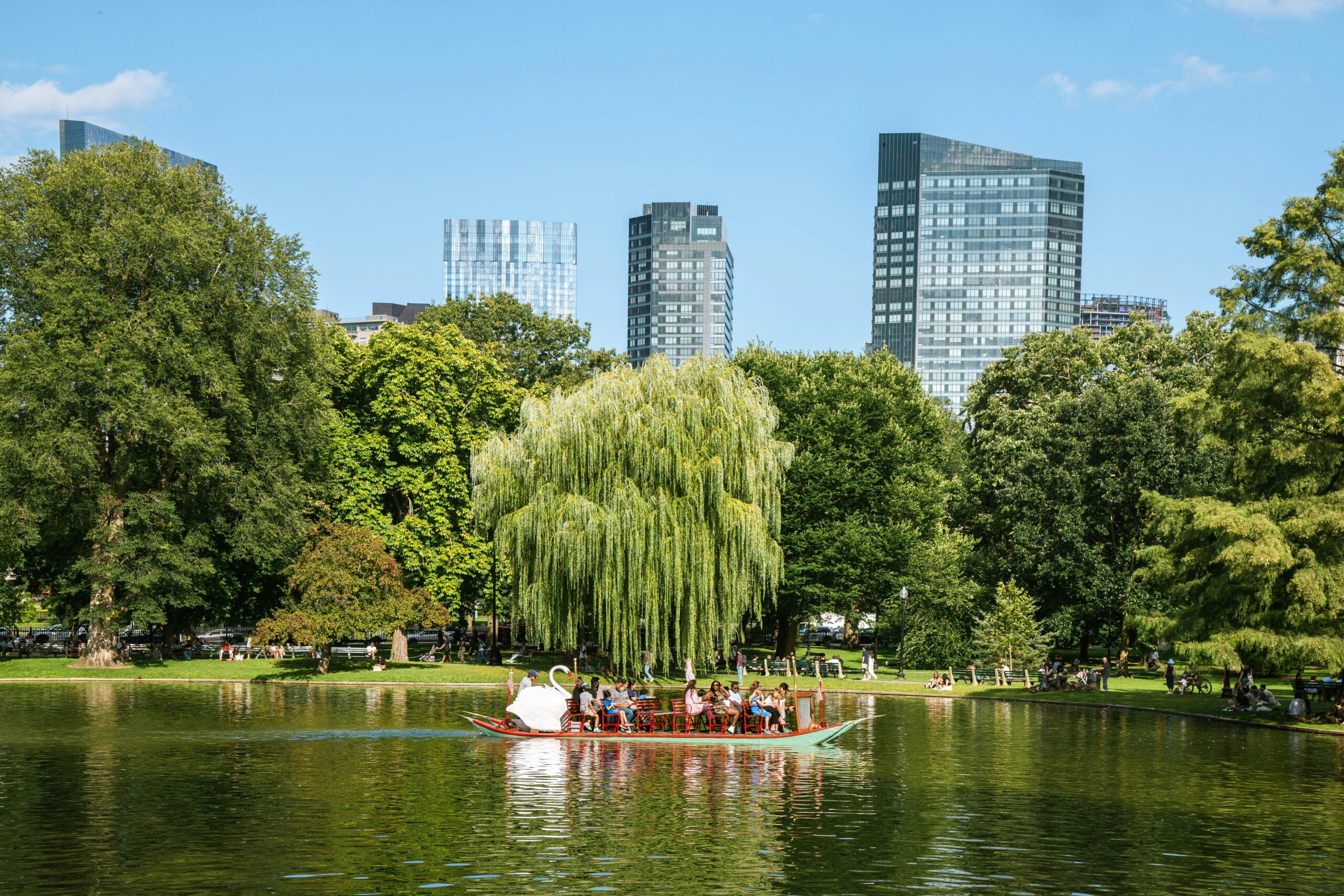 A swan-shaped paddleboat carries multiple passengers across a calm pond surrounded by lush green parkland with a prominent weeping willow tree in the center and various other mature trees. Modern glass high-rise buildings rise above the tree line in the background, while people can be seen enjoying the grassy areas around the water's edge under a blue sky with scattered white clouds.