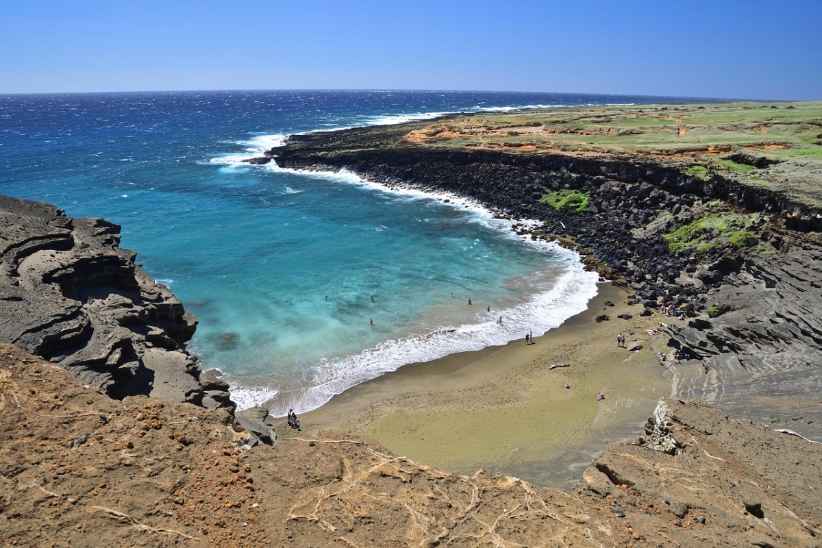 A crescent-shaped cove features olive-green sand surrounded by dark volcanic rock cliffs and formations, with turquoise water and white waves breaking along the shore. Several people are scattered across the beach, while the rocky coastline extends outward with grass-covered terrain visible on the higher ground under a clear blue sky.