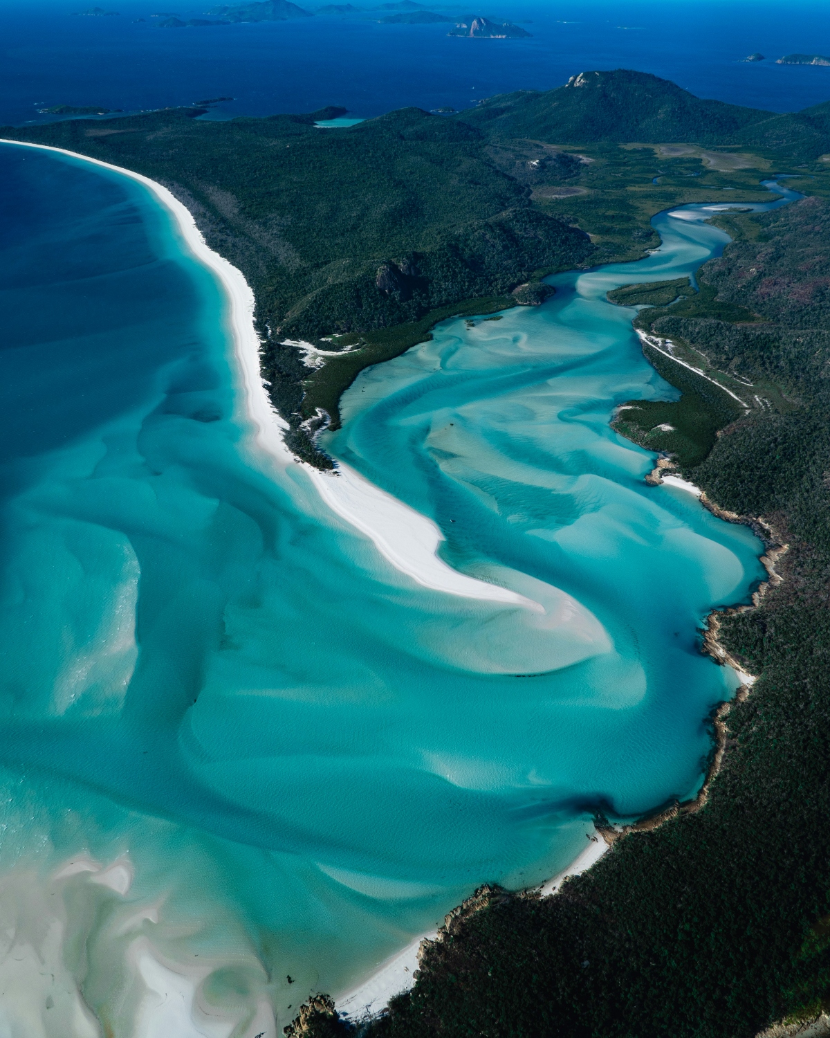 An aerial view shows a curved white sand beach bordered by densely forested hills, with a protected inlet displaying swirling patterns of turquoise and light blue water created by shallow sandbars and tidal movements. Multiple small islands are visible in the deeper blue ocean waters beyond the bay, while the pristine white sand creates striking contrast against the varying shades of blue water.
