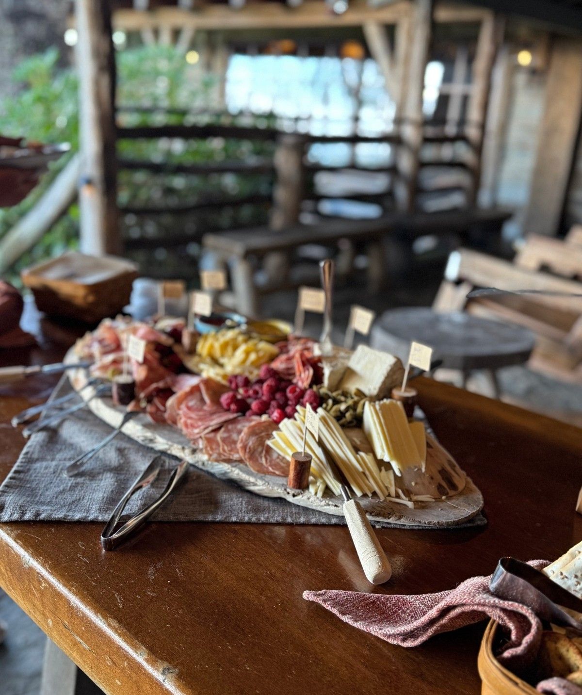 This image shows a charcuterie board arranged on a dark slate serving tray, featuring an assortment of cured meats, cheeses, crackers, and garnishes like berries and olives with small identification labels. The board is positioned on a wooden table surface with cheese knives and other utensils nearby, and a striped cloth napkin is visible to the side. The background shows a blurred restaurant or dining establishment interior with seating and greenery visible through windows.