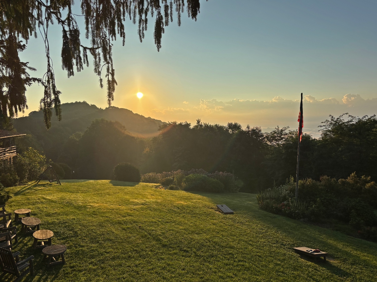 This image shows a large grassy lawn area during sunrise, with the sun visible in a hazy sky above rolling forested hills in the background. The scene includes outdoor furniture such as wooden picnic tables on the left side, a flagpole with a flag on the right, and trees including one with drooping branches in the upper left corner. 