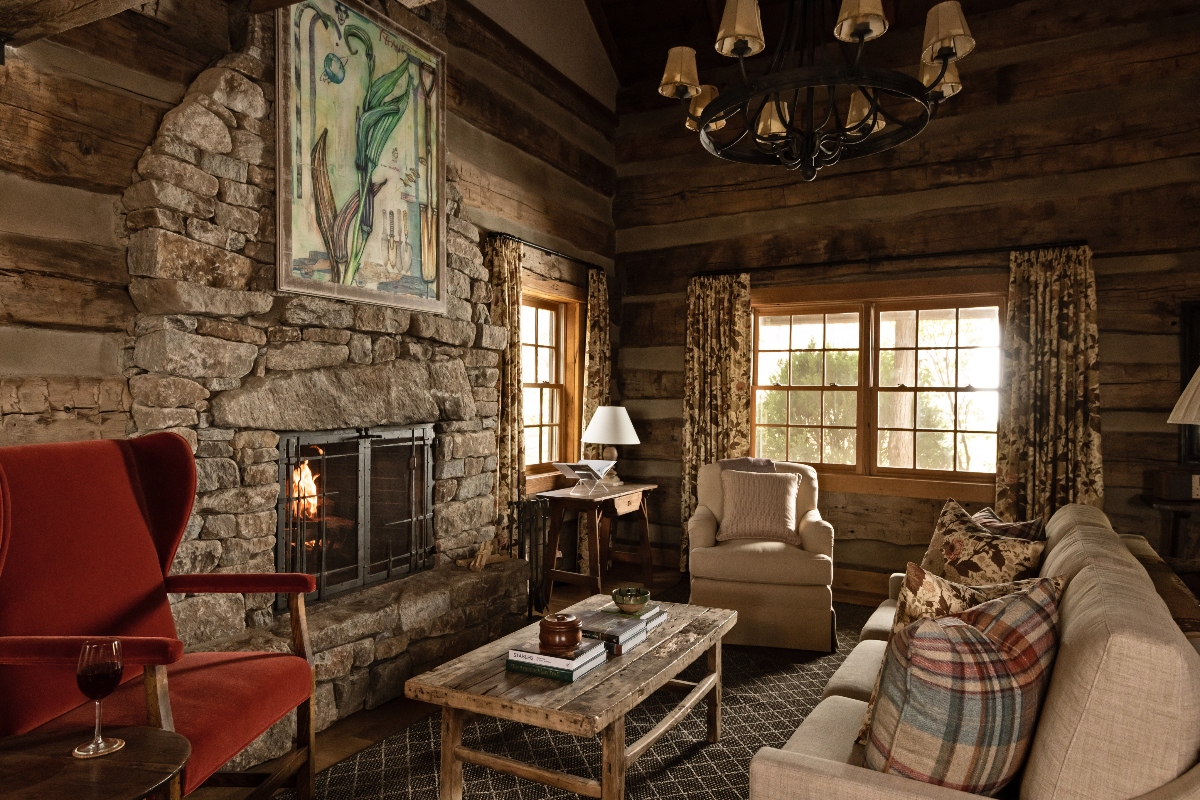This rustic interior features a stone fireplace with an active fire, flanked by a red upholstered chair and a beige sofa with plaid throw pillows. The room has log walls, multi-paned windows with patterned curtains, a wooden coffee table, and a wrought iron chandelier hanging from the exposed beam ceiling.