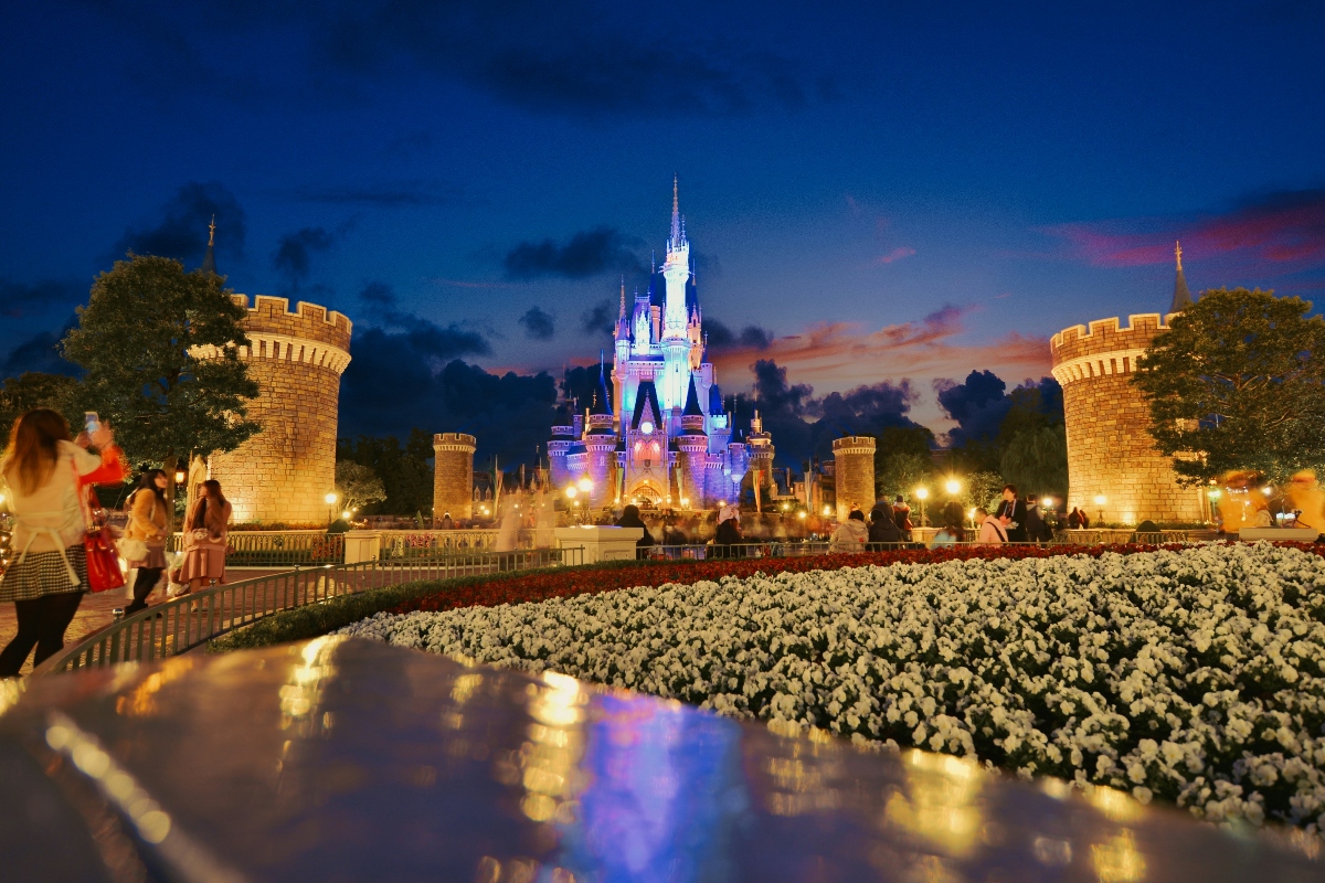 A fairytale castle illuminated with blue and purple lights stands at the center of the frame during twilight, with dramatic clouds in shades of blue, pink, and orange in the sky behind it. The castle is flanked by illuminated stone turrets and towers along a wide pathway, with beds of white flowers lining the walkway in the foreground. Visitors can be seen as blurred figures moving along the path, with trees framing both sides of the scene and warm lighting throughout the area.