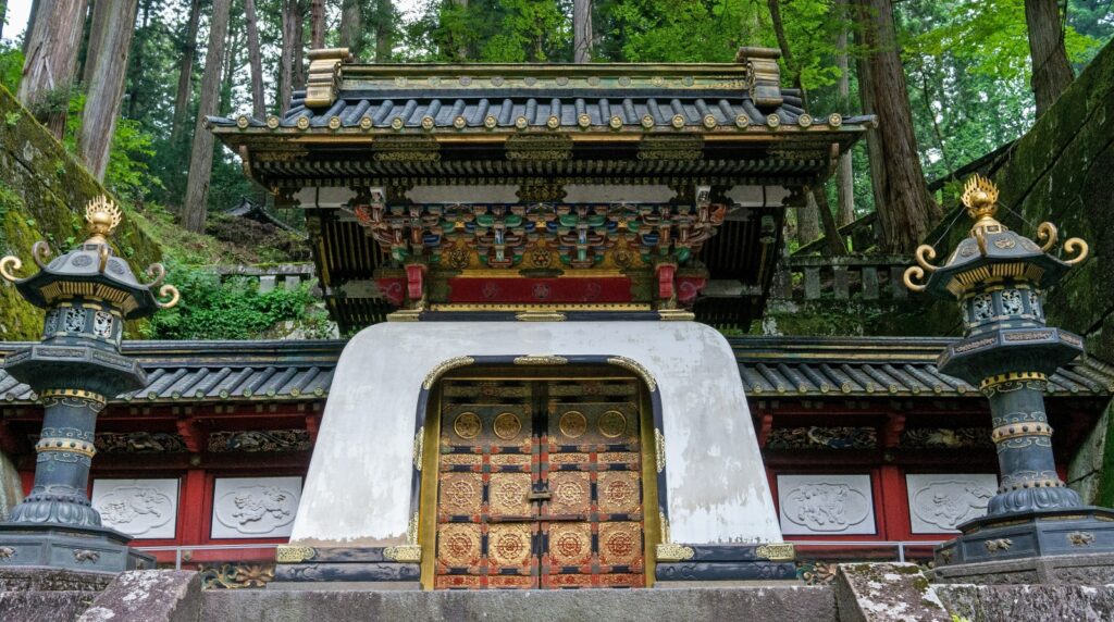 A traditional Japanese temple gate featuring ornate gilded double doors with intricate decorative patterns, framed by a distinctive white curved archway. The structure displays elaborate multi-tiered roofing with dark tiles and colorful carved woodwork in red, black, and gold, flanked by two ornamental bronze lanterns on decorative pedestals. The temple entrance is set within a dense forest of tall trees with vibrant green foliage, accessed by stone steps at the base.