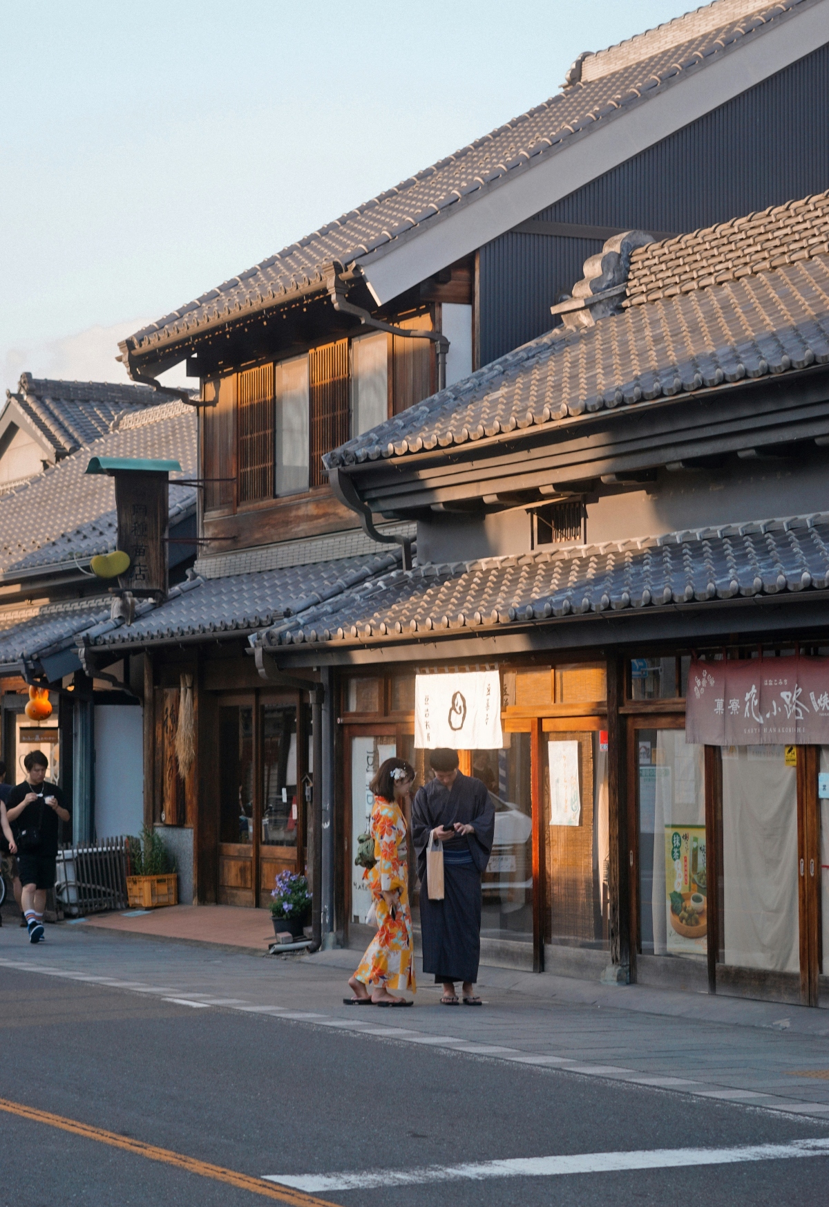 A traditional Japanese street featuring historic buildings with layered curved tile roofs and dark wooden facades during golden hour. Two people wearing colorful kimono stand in front of illuminated storefronts with traditional sliding doors and white fabric noren curtains. The architecture displays characteristic elements of Japanese design including multiple overlapping rooflines and wooden lattice details.