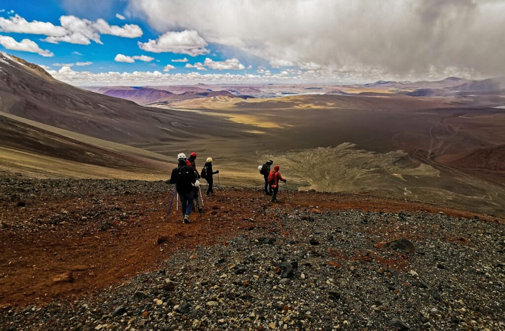 A group of hikers with backpacks and trekking poles stands on a rocky, barren slope overlooking an expansive arid landscape of rolling hills and distant mountains. The desert terrain features brown and tan-colored earth with sparse vegetation under a dramatic sky filled with white clouds against a bright blue background.