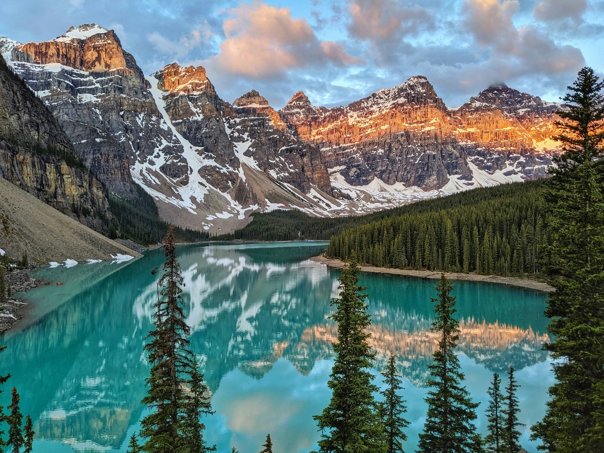 A pristine turquoise lake reflects snow-capped mountain peaks with distinctive layered rock formations illuminated by warm golden light, surrounded by dense evergreen forest. The calm water surface creates a perfect mirror image of the dramatic mountain range and cloudy sky, with tall coniferous trees framing the foreground of the alpine scene.