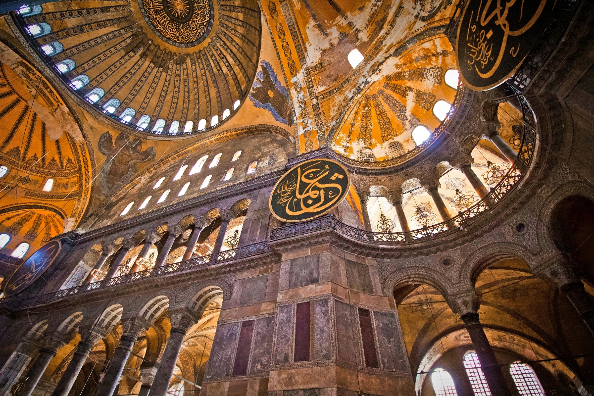 The interior view shows a massive domed ceiling with intricate patterns and a ring of windows around its base, supported by ornate arches and multiple levels of columned galleries. Large circular medallions with Arabic calligraphy are prominently displayed on the walls and pillars, while warm golden lighting illuminates the elaborate Byzantine architectural details throughout the vast space.